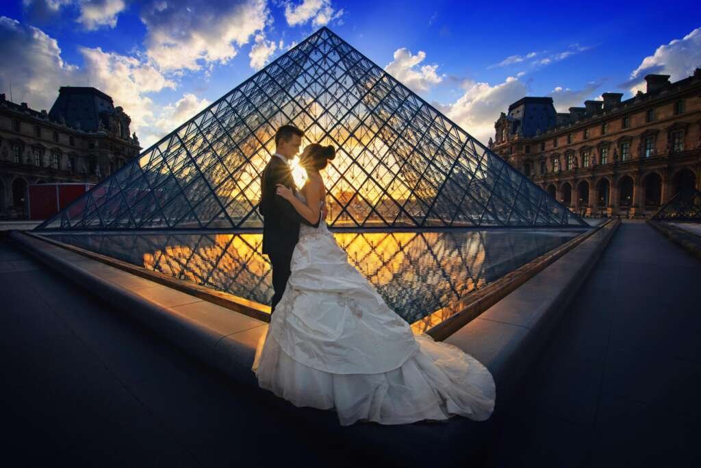 Couple embraces before the Louvre Pyramid during a stunning sunset in Paris.